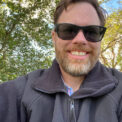 Selfie photo of a middle-aged caucasian man with a beard at a farm wearing dark sunglasses, a black jacket sitting under green trees.